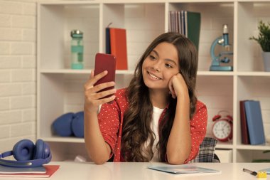 Mutlu kıvırcık saçlı çocuk sınıfta telefonda selfie çekiyor, video kaydediyor.