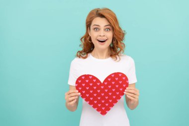 Şaşırmış kızıl saçlı kadın mavi arka planda kırmızı kalbi tutuyor..