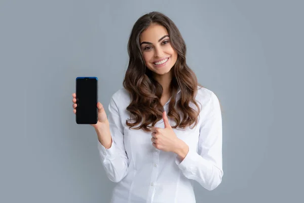 Mutlu genç kadın boş akıllı telefon ekranı gösteriyor. Tatmin olmuş, gülümseyen iş kadını cep telefonuna mesaj yazıyor, çevrimiçi iletişimden hoşlanıyor, geri bildirim türleri, gri stüdyo duvarında izole edilmiş.
