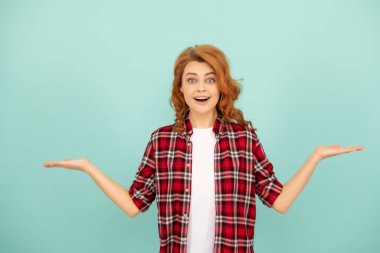 Kareli tişörtlü kızıl saçlı kadın mavi arka planda ürün sunuyor..