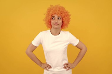 Yukarı bakıyorum. Turuncu peruklu komik kız. 1 Nisan şakası. Mutlu bayan eğleniyor. Deliriyor. Gülümseyen süslü parti bakışı. Sarı arka planda palyaço peruğu takan ucube kadın. insan duygularını ifade etmek.