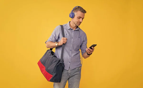 man listen music and hold sports bag. modern life and lifestyle. guy chatting in earphones. mature businessman in headphones use smartphone with travel bag.