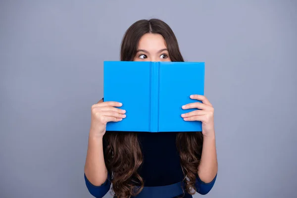 Schoolchild, teenage student girl hold book near eyes on gray isolated studio background. School and education concept. Back to school. Face hidden with book