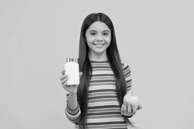 Mutlu kız elmayı ve takviye şişesini tutar sarı arka plan, vitaminler.