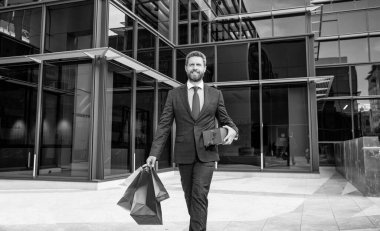 Resmi takım elbiseli, neşeli, başarılı sakallı iş adamı dışarıda alışveriş torbalarıyla dolaşıyor..