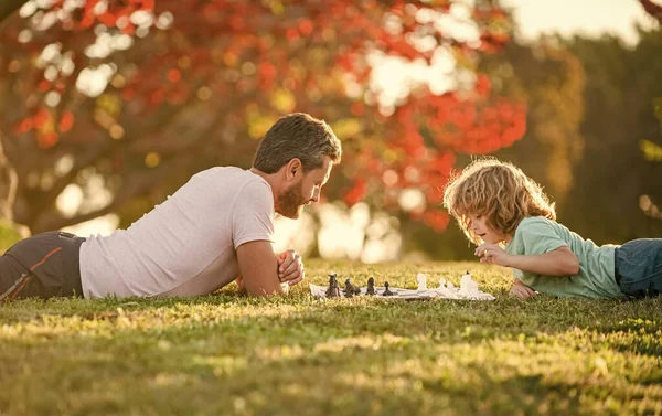 happy family of daddy and son child playing chess on green grass in park outdoor, erudite.