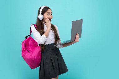 Okula dönelim. Okul üniformalı, çantalı, kulaklıklı ve dizüstü bilgisayarı olan mutlu bir kız öğrenci. İzole edilmiş mavi arka planda okul çocukları. Heyecanlı bir yüz. Şaşırtıcı ifade.