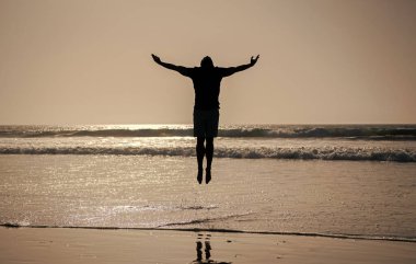 Yazın plajda kaldırılmış ellerle atlayan atletik bir adamın silüeti..