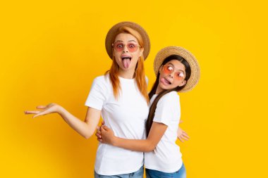 Annesiyle komik çocuğun stüdyo portresi. Komik surat. Anne ve genç kız sarılıyor, beyaz tişörtler giyiyor hasır şapka ve güneş gözlüğü takıyor, parlak sarı arka planda izole edilmiş.