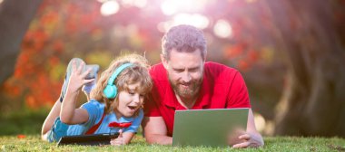 Baba oğul okul çocuğu, dizüstü bilgisayar eğitimi almış çimlerin üzerinde yatıyor, mutlu baba oğul ailesi parkta dizüstü bilgisayar ve kulaklık kullanıyor, mutlu aile.