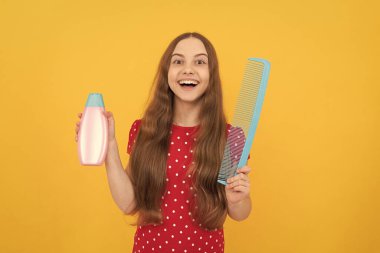Şaşırmış çocuk, sarı arka planda uzun saçlar için şampuan ve saç fırçası sunuyor..