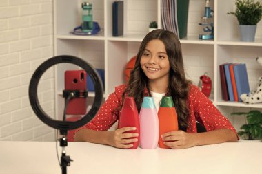 Şampuan şişeli vlogger. Akıllı telefondan video blogu hazırlıyorum. Blog lambası. Saç kremi. Neşeli kız selfie çekti. Çocuk güzellik blogcusu. Gençler için kozmetik ürünü sunan çocuk.