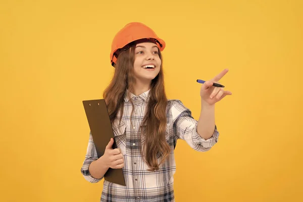 happy labour day. future career. teen girl in helmet making notes. builder kid write in folder. building and construction. happy child worker wear hardhat hold clipboard. pointing finger. childhood.