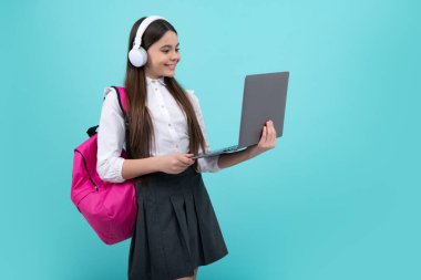 Okula dönelim. Okul üniformalı, çantalı, kulaklıklı ve dizüstü bilgisayarı olan bir kız öğrenci. İzole edilmiş mavi arka planda okul çocukları