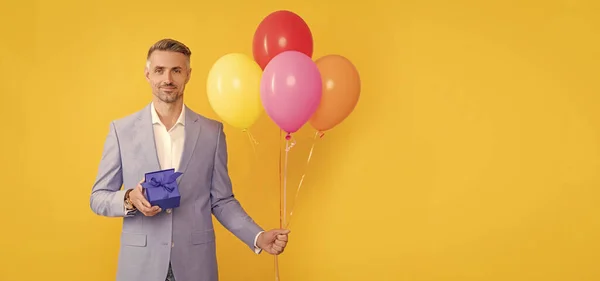 happy mature man with box and party balloons on yellow background, copy space, mens day.
