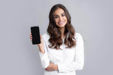 Mutlu genç kadın boş akıllı telefon ekranı gösteriyor. Genç bir kadın cep telefonundan mesaj okuyor ve gülümsüyor. Akıllı telefonlu mutlu iş kadını, gri arka planda duruyor.