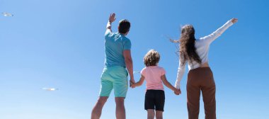 Deniz kıyısında kucaklaşan aile bayrağı. Yaz tatili, yatay afiş. Anne, baba ve çocuk gelecekte el ele tutuşacaklar. Yazın, arka planda, hayırseverlikte.