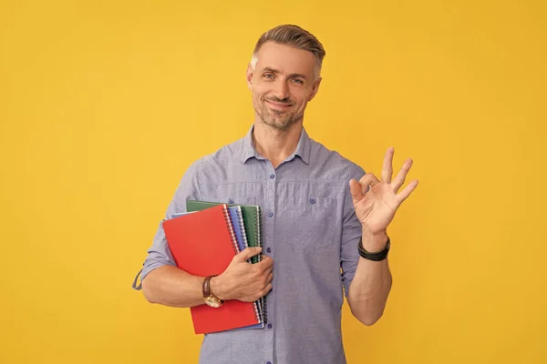 school teacher or university tutor. happy man hold folder showing ok gesture. mature man on yellow background with project. guy with documents in clipboard. signing a contract.