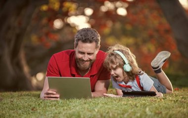 Bilgisayarda online eğitim. Baba ve oğul parkta modern iletişim teknolojisini kullanıyorlar. Mutlu aile blogu. Çocukluk ve ebeveynlik. Genç çocuk kulaklıkla müzik dinliyor. webinar video dersi.