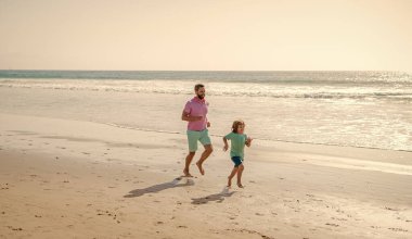 Hafta sonu aile günü. Baba ve çocuk dışarıda eğleniyor. Baba oğul güneşli yaz plajlarında koşuyorlar. Aile seyahati ve tatil. Çocukluk ve ebeveynlik. Babası denizdeki ya da okyanustaki çocukla.