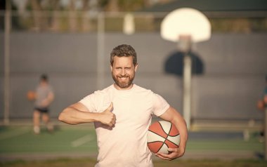 Neşeli kaslı erkek sporcu basketbol oyuncusu topu açık havada tutuyor başparmak hareketi ve spor başarısı gösteriyor..