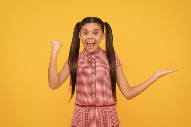 surprised teen girl presenting product on yellow background copy space celebrate success, promotion.