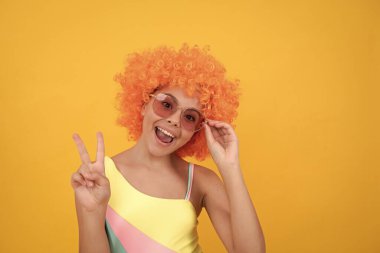 Güneş gözlüklü ve mayo giyen mutlu çocuk turuncu kıvırcık saçlı barış jesti yapıyor..