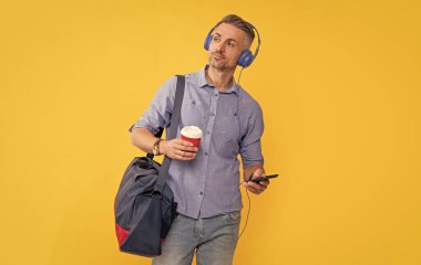 Modern hayat ve yaşam tarzı. Adam kahve içerken kulaklıkla sohbet ediyor. Kulaklık takan yetişkin bir iş adamı seyahat çantasıyla akıllı telefon kullanır. Müziği dinle ve spor çantasını tut..