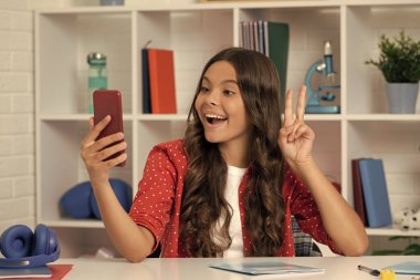 Şaşırmış çocuk uzun kıvırcık saçlı, akıllı telefonuyla selfie çekiyor barış jestleri yapıyor, blog yazıyor..