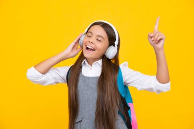 School leisure. Amazed child singing. School girl, teenager student in headphones on yellow isolated studio background. School and music education concept. Back to school