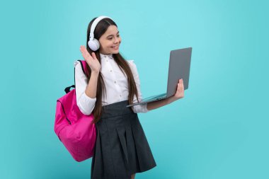 Okula dönelim. Okul üniformalı, çantalı, kulaklıklı ve dizüstü bilgisayarı olan bir kız öğrenci. İzole edilmiş mavi arka planda okul çocukları