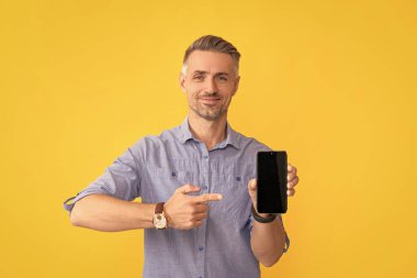 Gülümseyen adam sarı arkaplanda telefonu işaret ediyor, kopyalama alanı, sunum.