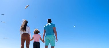 Family of mom father and child looking in future hold hands in summer, back view, summer vacation. Banner of family embracing on sea beach. Summer family vacation, horizontal banner