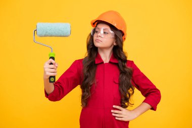 Çocukların tadilatı ve inşaat konsepti. Sarı arka planda izole edilmiş duvarları boyamak için koruyucu miğferi ve boya fırçası olan küçük bir kız.