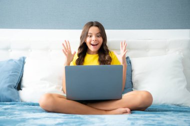 Teenager child girl browsing internet in a laptop in the bedroom at home. Expressive emotional excited teen girl