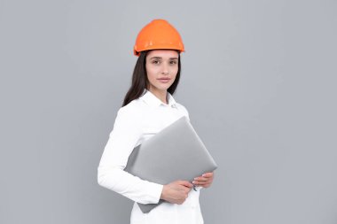 Gri arka planda dizüstü bilgisayarı olan kasklı genç bir kadın var. Mimar kadın kask ve gömlek giyer. İş kadını, inşaat müdürü, inşaatçı.