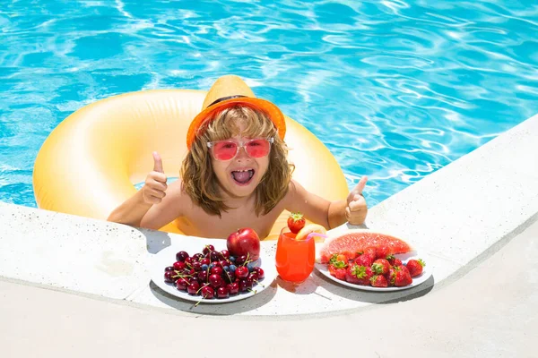 Happy child playing in swimming pool. Summer kids vacation. Summer fruit for children. Little kid boy relaxing in a pool having fun during summer vacation