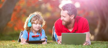 İnternette çim, çocukluk ve ebeveynlik üzerine uzanan dizüstü bilgisayarı olan bir okul çocuğuna baba pankartı. Mutlu çocuk kulaklıkla müzik dinler. webinar video dersi.