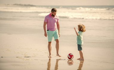 Baba ve çocuk dışarıda eğleniyorlar. Çocukluk ve ebeveynlik. Aile tatilleri. Spor aktivitesi. Baba ve oğul plajda futbol oynuyorlar. Güneşli bir günde babasıyla küçük oğlu. hafta sonu aile günü.