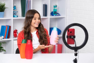 Çocuk video kaydedicisi telefonda video blogu yapıyor. Web günlüğü ve vlog. Genç kız blogger etkileyicisi sosyal medya için saç kremi ya da şampuan şişesi tutuyor. Mutlu kız yüzü, pozitif ve gülümsüyor.