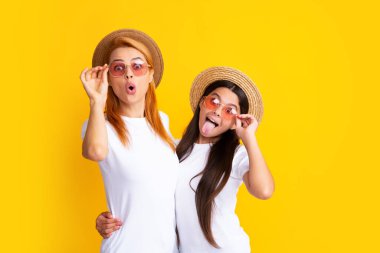 Studio portrait of funny child with mum. Funny face. Mom and teenager daughter hugging lovely cuddling, wearing white t-shirts straw hat and sunglasses, isolated on bright yellow background