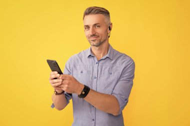 Modern kablosuz kulaklıklarda gülümseyen olgun bir adam sarı arka planda akıllı telefon ve iletişim kullanıyor..
