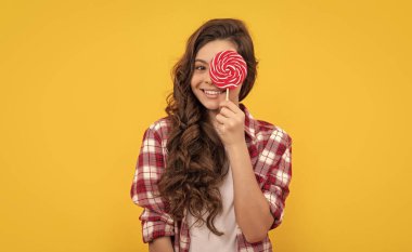 Uzun kıvırcık saçlı, kareli tişörtlü komik çocuk sarı arka planda lolipop karamel şekeri tutuyor, nefis..