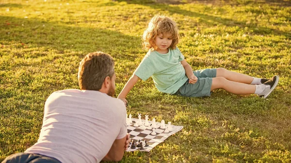happy family of daddy and son boy playing chess on green grass in park outdoor, parenthood.
