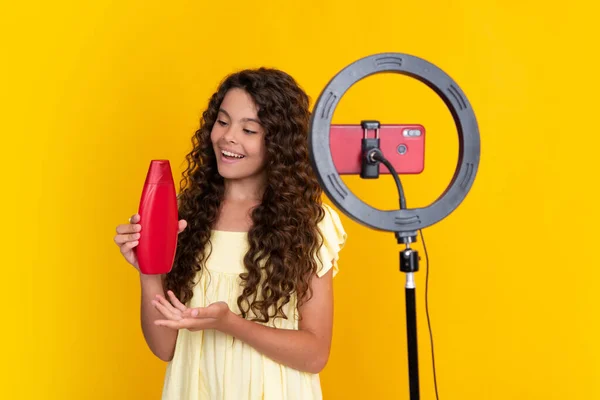 Teenager blogger making video hold hair conditioner or shampoo bottle, child vlogger influencer record content. Child blogger records video on mobile phone using ring video lamp