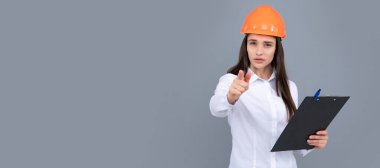 Gri arka planda panosu olan koruyucu kasklı ciddi bir kadın. Genç kadın inşaat müdürü. Kasklı kadın ve kadın işçi seni işaret ediyor.
