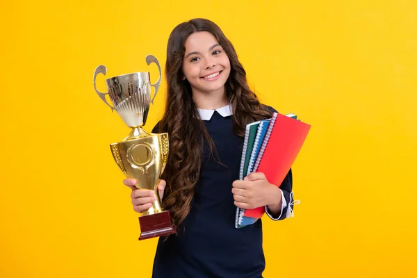 Teenager school girl with award winner trophy. Child hold books with gold trophy or winning cup isolated on yellow. Education graduation, victory and winning
