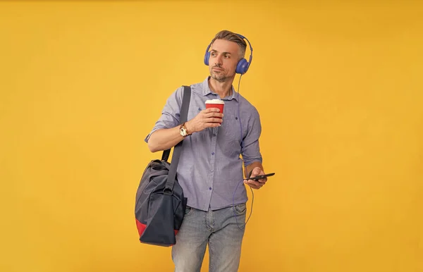 guy listen music in headphones chat on smartphone with travel bag drinking coffee on yellow background, modern life.