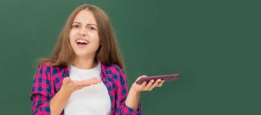 confused child at school chatting on phone on blackboard background. Portrait of schoolgirl student, studio banner header. School child face, copyspace