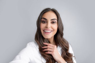 Pleasant girl making selfie in studio and laughing. Good-looking young woman with brown wavy hair taking picture of herself on gray background